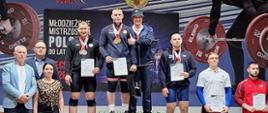Zawodnicy stoją na podium podczas ceremonii dekoracji na Młodzieżowych Mistrzostwach Polski w podnoszeniu ciężarów. W tle widoczny baner zawodów, a przed podium sztanga z obciążeniami.