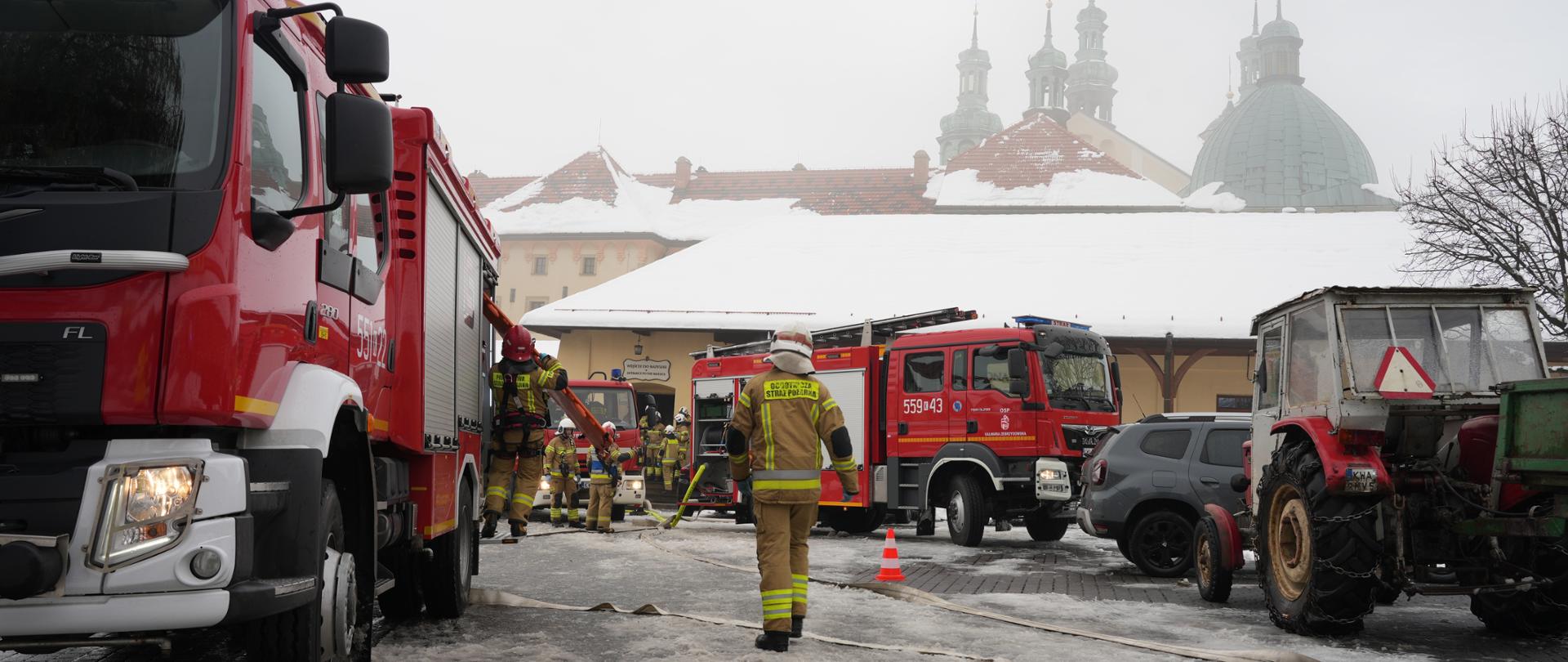 Zdjęcie przedstawia strażaków prowadzących działania podczas ćwiczeń na terenie klasztoru w Kalwarii Zebrzydowskiej. Na pierwszym planie widać kilka wozów strażackich ustawionych na zaśnieżonym placu, strażaków w pełnym umundurowaniu oraz rozwinięte węże gaśnicze. W tle widoczne są zabudowania klasztorne z ośnieżonym dachem i kopułami kościoła, częściowo spowite mgłą. Atmosfera wskazuje na zimowe warunki i intensywną pracę służb.