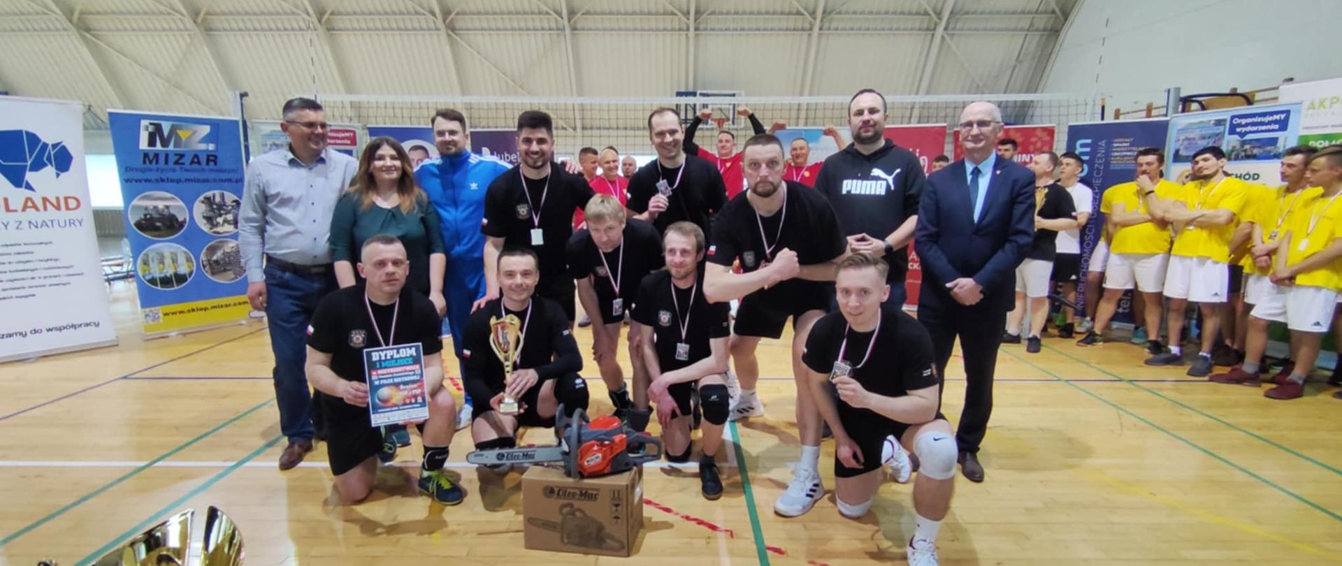 Wygrana drużyna z Ochotniczej Straży Pożarnej w Kraśniku w czarnych koszulkach i spodenkach. czterech zawodników na pierwszym planie przyklęka na jedno kolano, zaś pozostali stoją tuż za nimi. Zawodnicy prezentują na szyjach medale, a przed nimi na podłodze stoi ustawiona nagroda: piła elektryczna. Z prawej strony stoją: Starosta Kraśnicki Zbigniew Dżugaj, a obok niego Wójt Gminy Trzydnik Duży Krzysztof Serafin. Jako trzeci od lewej stoi Prezes Stowarzyszenia Wschód Lubelskich Serc Michał Stawiarski.