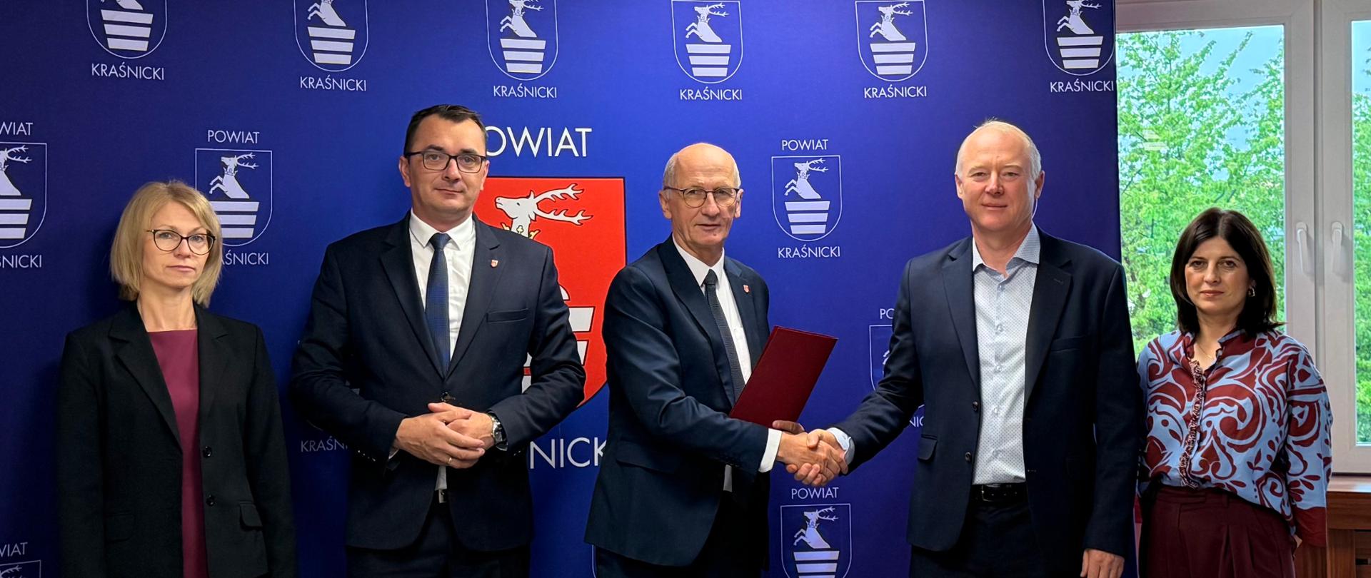 Pamiątkowe zdjęcie z podpisania umowy na wykonanie prac budowlanych. Na zdjęciu za stołem prezydialnym stoi pięć osób, które pozują do pamiątkowego zdjęcia z egzemplarzami podpisanej umowy. W tle znajduje się niebieska ścianka z wizerunkami herbu powiatu kraśnickiego.