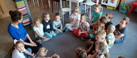 Dzieci siedzą na podłodze w kółku, razem z nimi prowadząca zajęcia