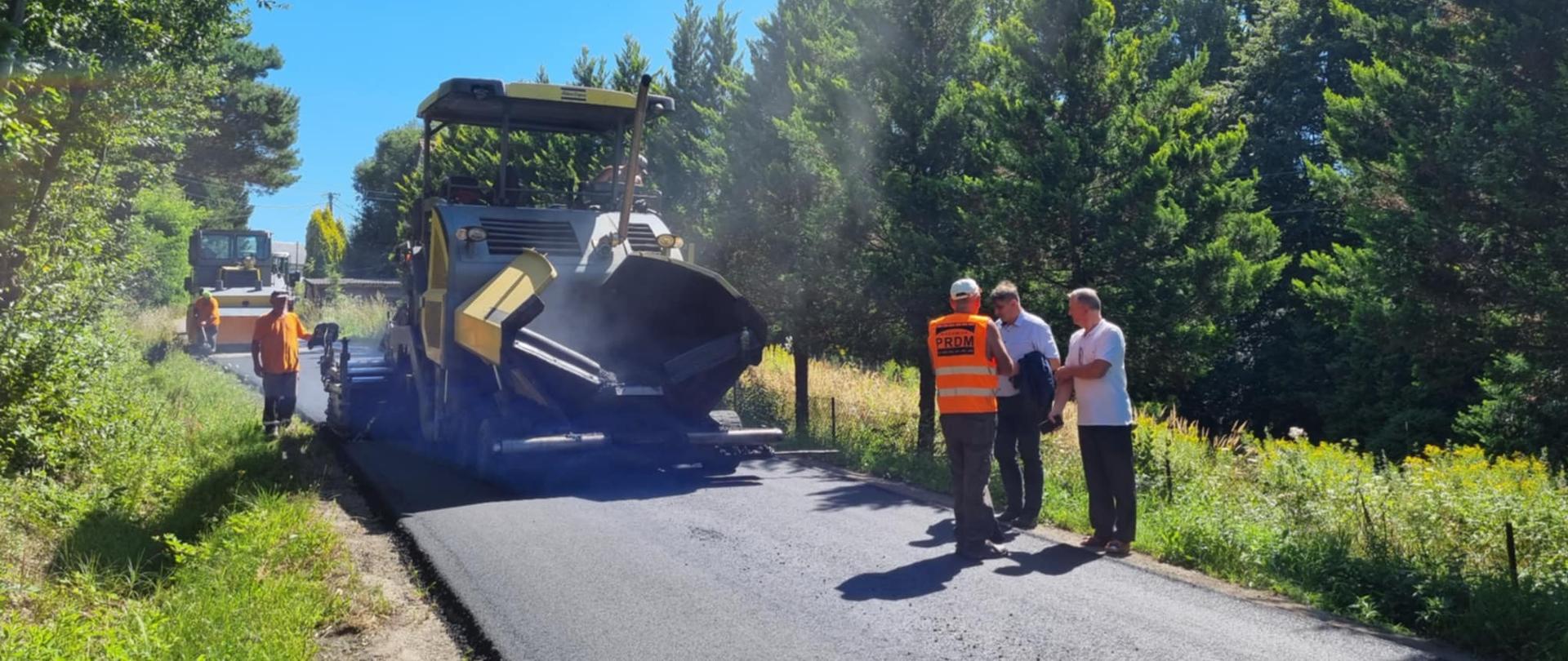 Na zdjęciu widać proces asfaltowania drogi w otoczeniu zielonych drzew. Na pierwszym planie znajduje się duża maszyna drogowa układająca nową warstwę asfaltu. Po lewej stronie pracuje dwóch robotników w pomarańczowych kamizelkach, a w tle widoczny jest kolejny pojazd budowlany. Po prawej stronie, przy krawędzi drogi, stoją trzy osoby rozmawiające ze sobą, z których jedna ma na sobie odblaskową kamizelkę z napisem „PRDM”. Cała scena odbywa się w słoneczny dzień pod bezchmurnym, niebieskim niebem.
