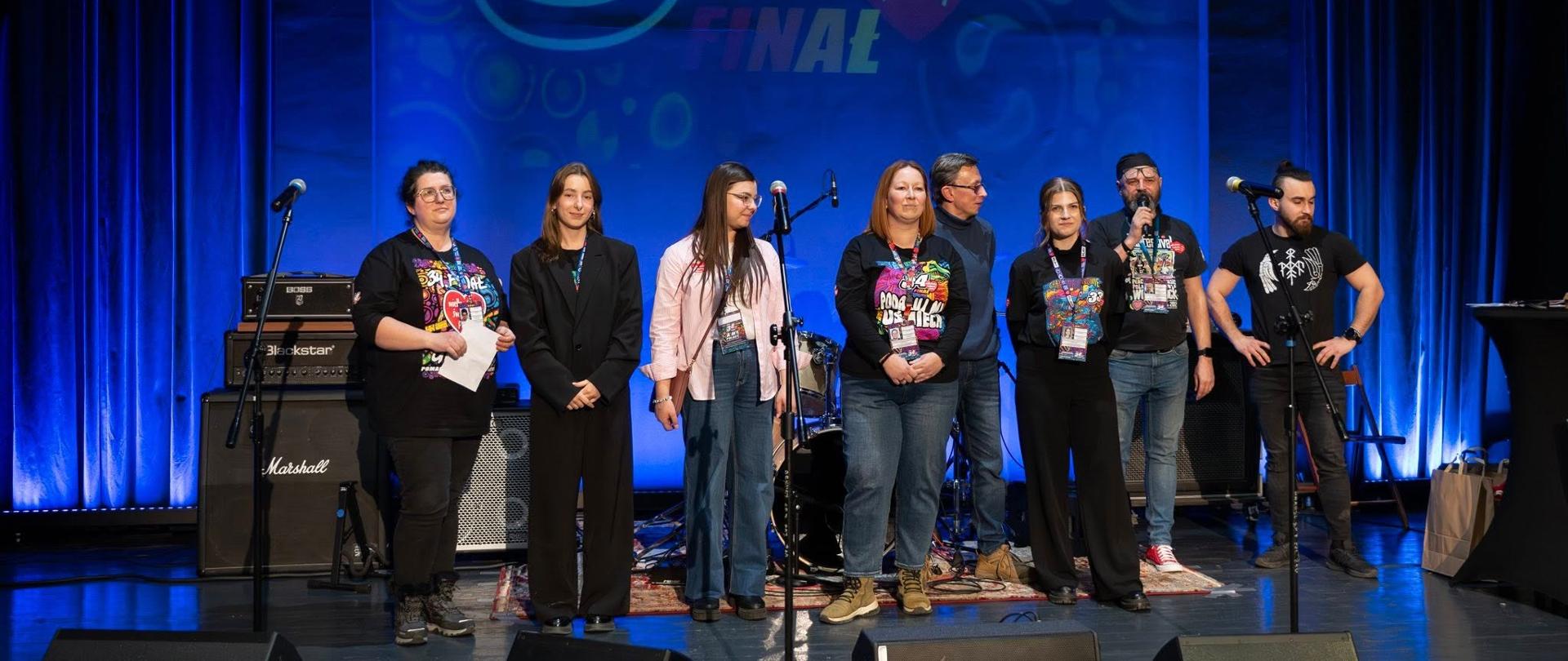 Na scenie stoi grupa wolontariuszy i organizatorów 34. Finału Wielkiej Orkiestry Świątecznej Pomocy 2026. W tle widoczne jest duże logo „34 Finał” z sercem WOŚP. Uczestnicy ustawieni są w rzędzie, obok mikrofonów i sprzętu muzycznego, w sali koncertowej o niebieskim oświetleniu.