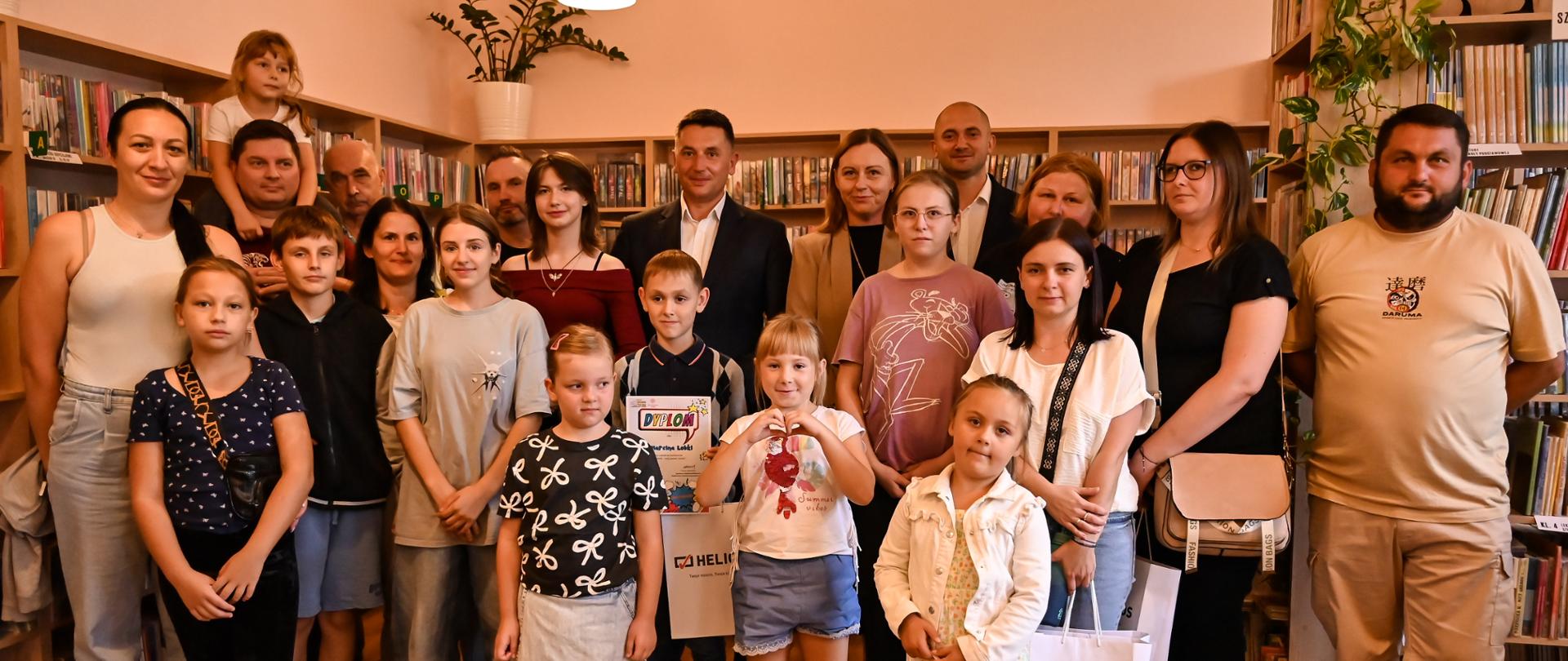 Zdjęcie grupowe nagrodzonych uczestników wraz z rodzicami i opiekunami oraz burmistrzem i zastępcą burmistrza. W tle biblioteczne regały z książkami.