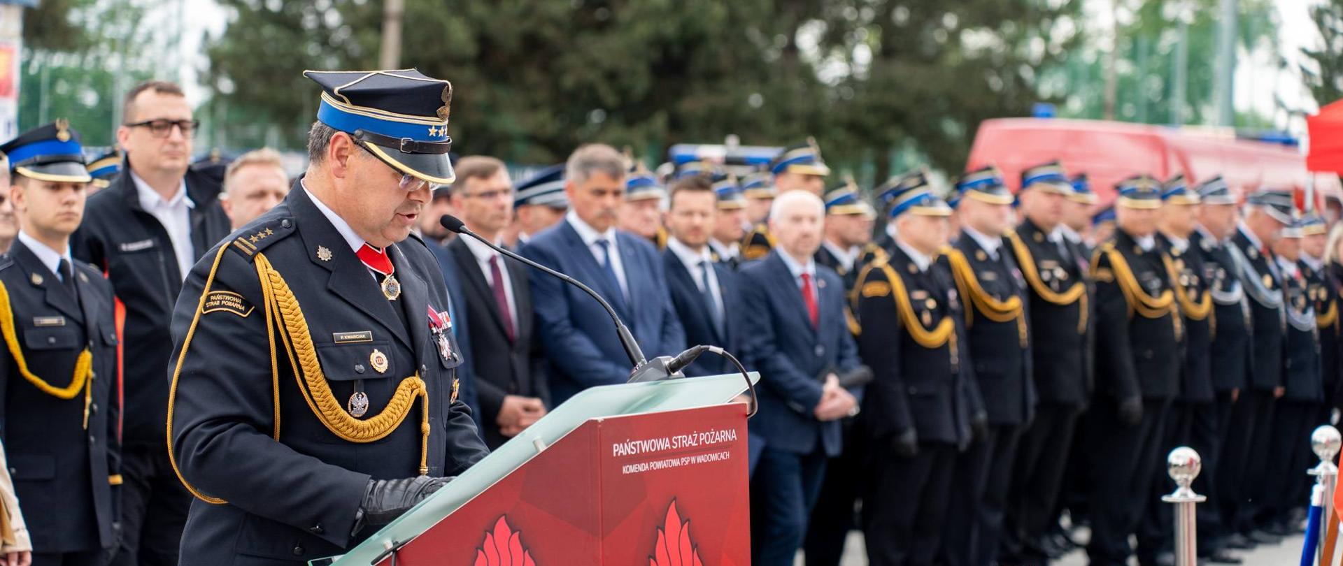 Komendant Powiatowy Państwowej Straży Pożarnej w Wadowicach przemawia przy mównicy podczas Powiatowych Obchodów Dnia Strażaka. W tle stoją strażacy w galowych mundurach oraz zaproszeni goście, w tym przedstawiciele samorządów i służb. Na pierwszym planie widoczne są czerwone róże, a uroczystość odbywa się na placu przed Komendą Powiatową PSP w Wadowicach.