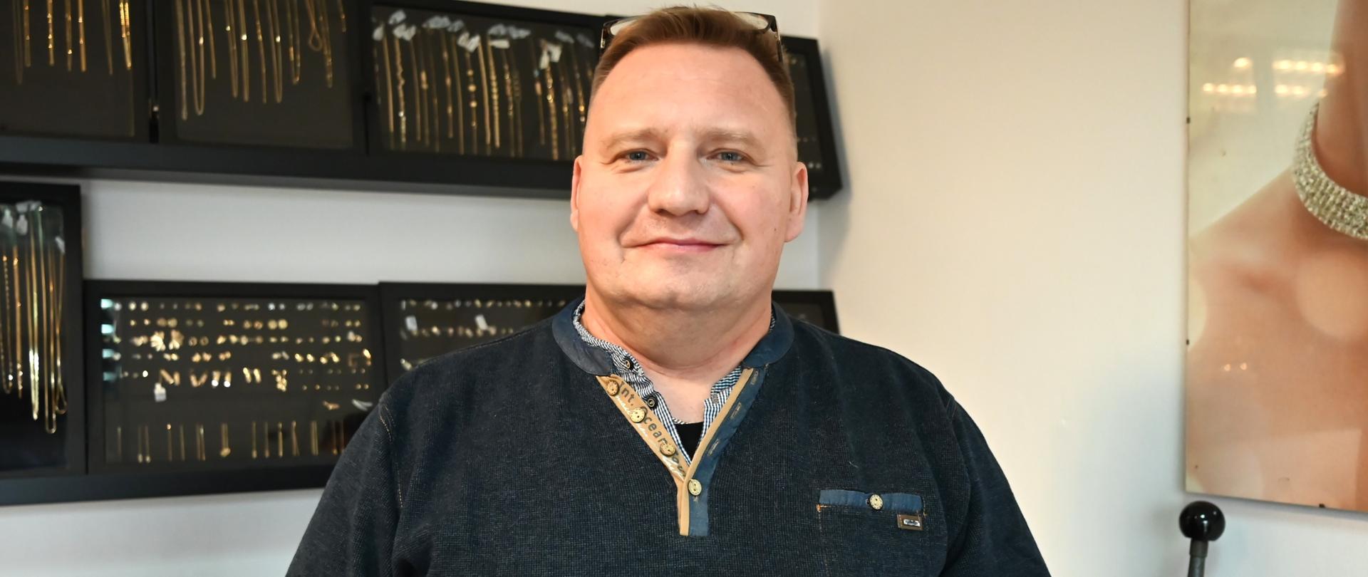 Uśmiechnięty mężczyzna stoi we wnętrzu sklepu jubilerskiego. Ma na sobie ciemny sweter i patrzy w stronę obiektywu. W dłoniach trzyma otwarte, czerwone pudełko jubilerskie, w którym znajduje się złota zawieszka w kształcie serca. Za nim, na ścianie, widoczne są czarne gabloty z wyeksponowanymi złotymi łańcuszkami i inną biżuterią. Oświetlenie jest jasne, a wnętrze sprawia wrażenie eleganckiego i profesjonalnego.