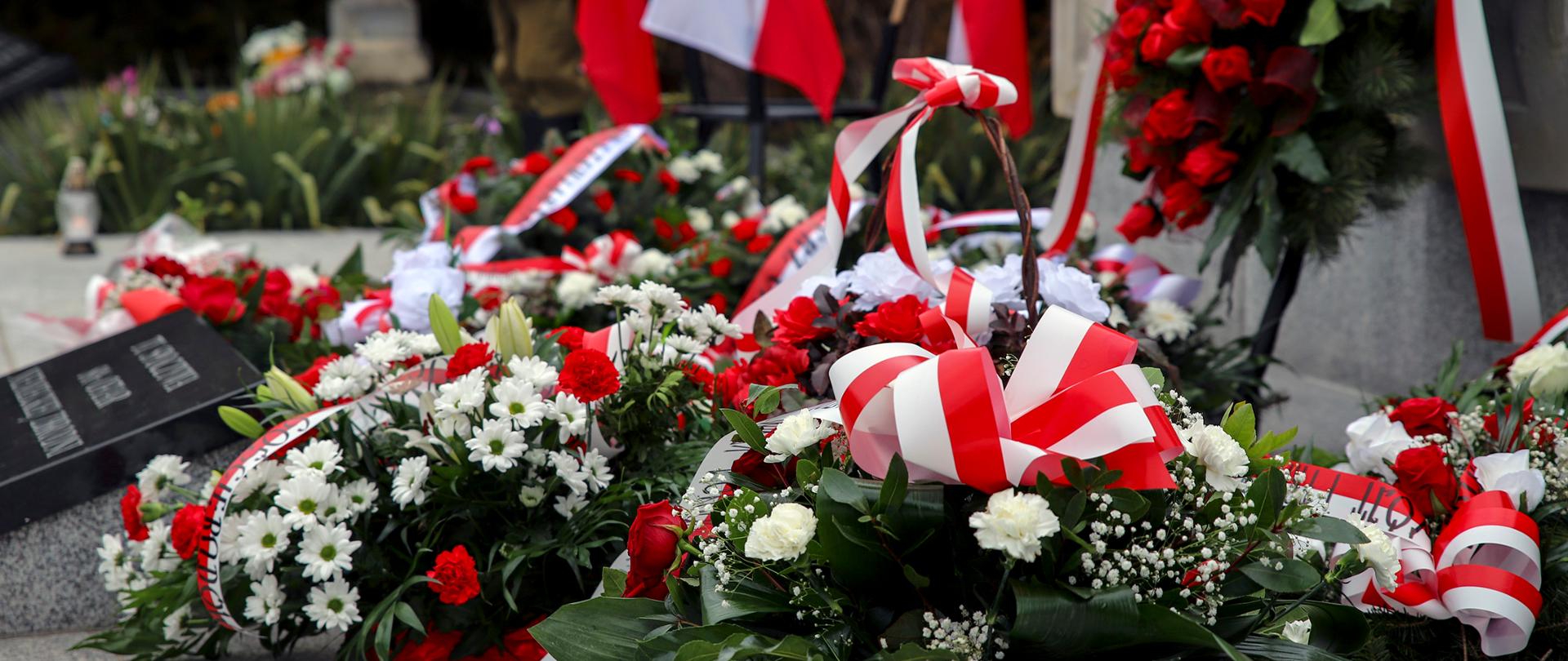 Na zdjęciu wiązanki biało-czerwonych kwiatów ozdobionych biało-czerwonymi wstęgami. Wiązanki zostały złożone przy pamiątkowej mogile na cmentarzu w Borowie. W tle znajdują się biało-czerwone flagi.