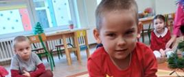 Dzieci siedzą w sali zabaw, na dywanie. W centrum zdjęcia widać chłopczyka, który siedzi przy choince, trzyma bombkę, wiszącą na drzewie