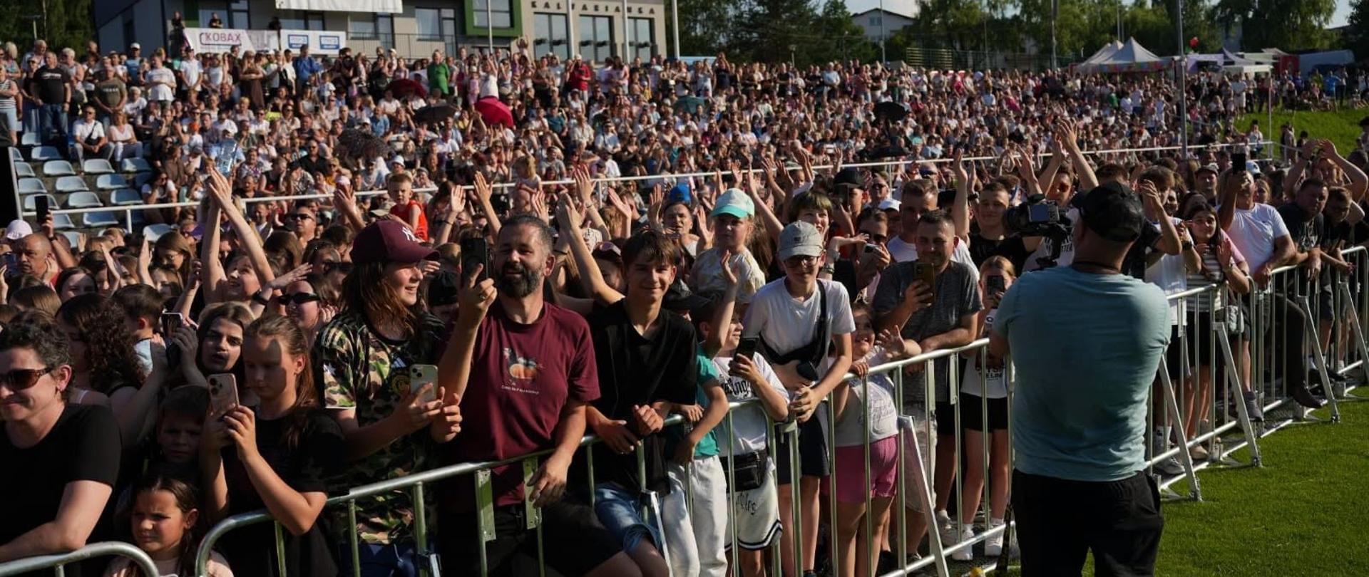 Licznie zgromadzona publiczność przed sceną plenerową na stadionie w Kalwarii Zebrzydowskiej podczas gminnego wydarzenia. Uczestnicy stoją przy barierkach i na trybunach, aktywnie uczestnicząc w koncercie.
