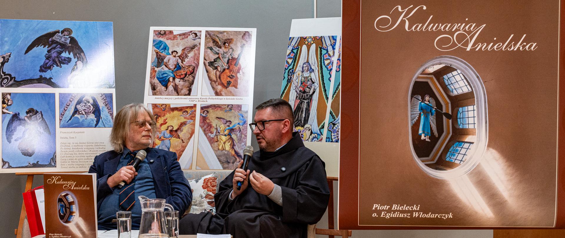 Dwóch mężczyzn siedzi na scenie podczas spotkania autorskiego i prowadzi rozmowę z użyciem mikrofonów. Po lewej stronie starszy mężczyzna w okularach, ubrany w marynarkę i krawat, słucha uważnie rozmówcy. Po prawej zakonnik w habicie gestykuluje, wyjaśniając coś w trakcie wypowiedzi. Przed nimi znajduje się niewielki stolik z dzbankiem wody, szklankami i egzemplarzami książki.
W tle widoczne są plansze z reprodukcjami aniołów oraz duża grafika okładki albumu „Kalwaria Anielska”. Na podłodze obok sceny stoi torba prezentowa. Całość ma charakter kameralnego wydarzenia kulturalnego, związanego z promocją publikacji o tematyce sakralnej i artystycznej.
