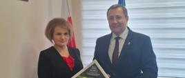 Kobieta z mężczyzną ubrani na galowo trzymając w ręku tabliczkę, w tle flaga Polski i Gminy Raba Wyżna