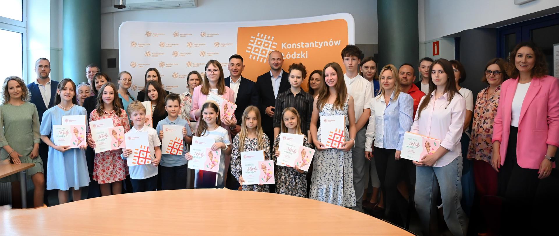 Grupowe zdjęcie uczestników i organizatorów uroczystości w Urzędzie Miejskim w Konstantynowie Łódzkim
Grupa dzieci, młodzieży i dorosłych pozuje do zdjęcia na tle ścianki z logo miasta Konstantynów Łódzki. Kilkoro dzieci trzyma w rękach dyplomy i teczki z nagrodami. W tle widoczne elementy sali konferencyjnej.