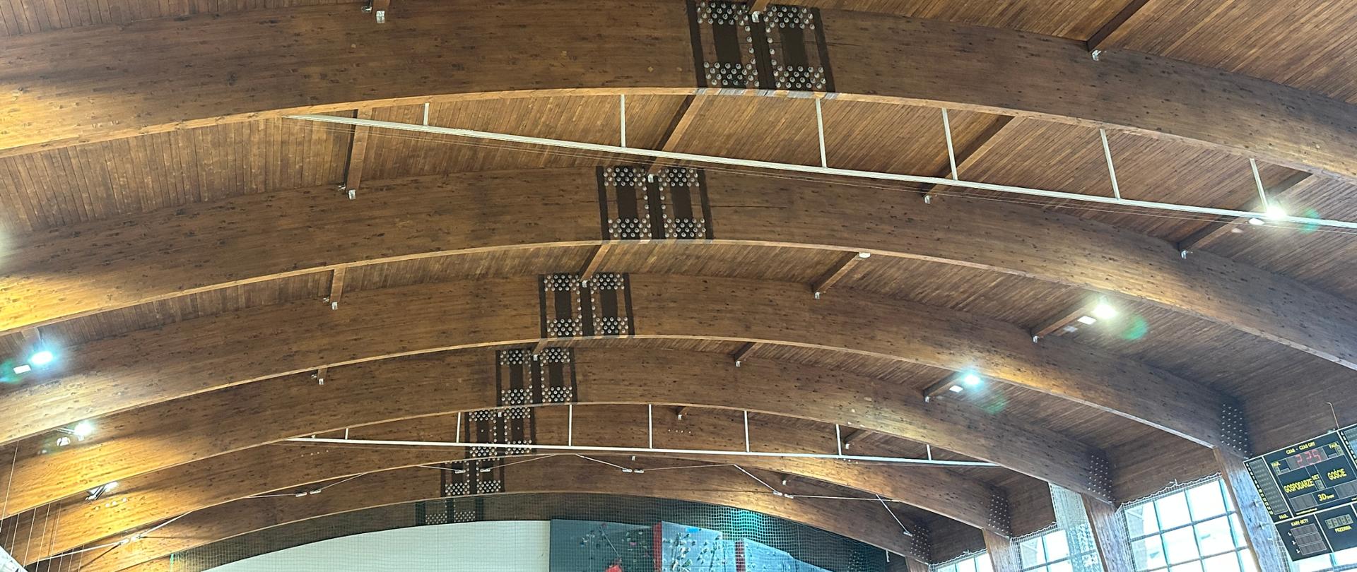 Wnętrze hali sportowej z widoczną drewnianą konstrukcją dachu opartą na masywnych, łukowych belkach. Na belkach zamocowane są metalowe łączniki i elementy wzmacniające, a między nimi biegną stalowe cięgna i instalacje. Oświetlenie sufitowe równomiernie doświetla przestrzeń. W tle widać fragment dużego okna oraz tablicy wyników sportowych.