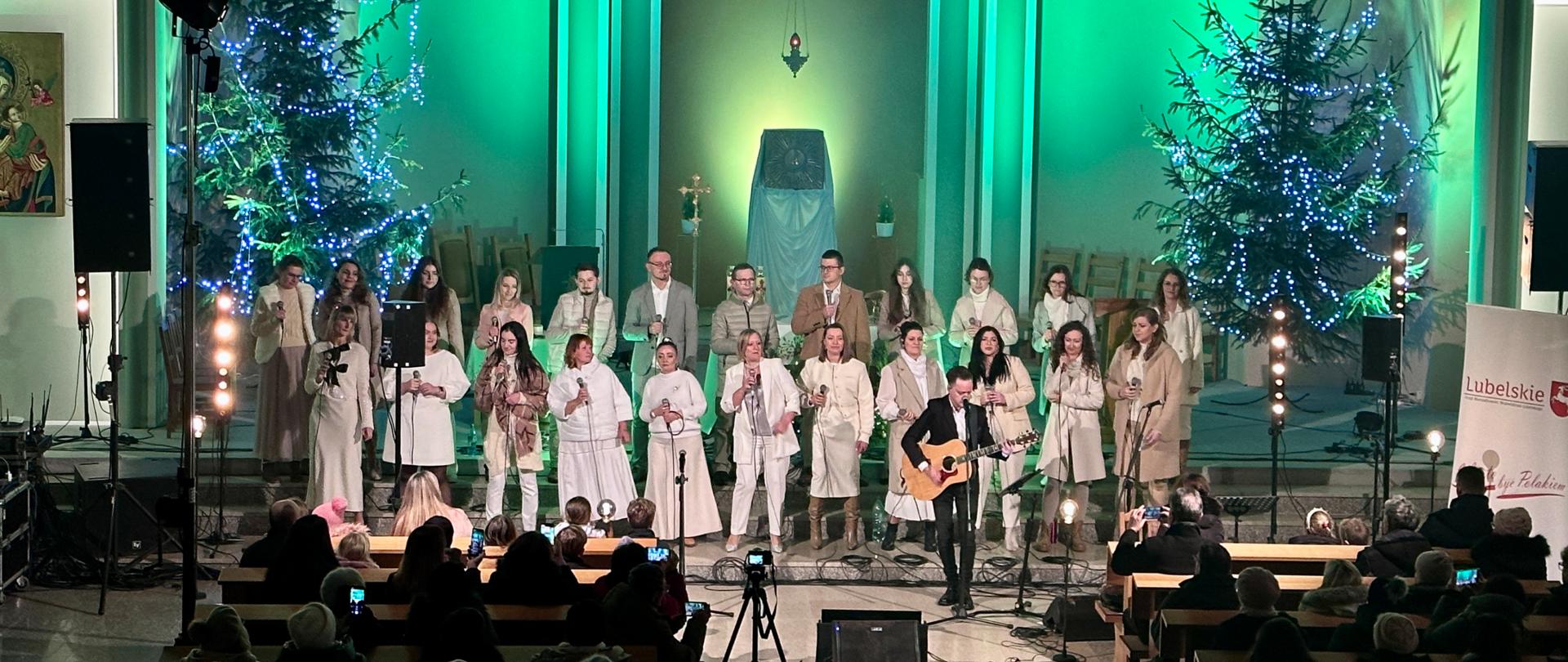 Zdjęcie z koncertu kolęd. na pierwszym planie znajdują się siedząca w kościelnych ławkach publiczność. Przed nimi ubrani w jasne stroje członkowie zespołu podczas występu. W tle znajdują się świąteczne choinki oraz oświetlony na zielono ołtarz.