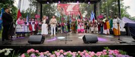 Na dużej scenie plenerowej stoją uczestnicy Międzynarodowego Festiwalu Folklorystycznego Polka w Konstantynowie Łódzkim. W centrum kobieta i mężczyzna w jasnych strojach zapowiadający koncert, wokół nich tancerze w barwnych strojach ludowych z różnych krajów świata, trzymający flagi narodowe oraz tabliczki z nazwami państw. Na pierwszym planie rabata z różowymi kwiatami.