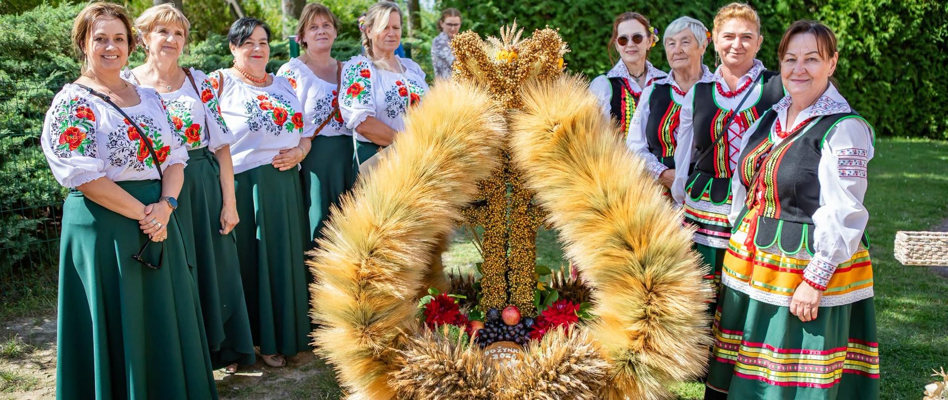 Zdjęcie przedstawiające dziewięć kobiet w strojach ludowych, stojących wokół dużego wieńca dożynkowego. Po lewej stronie pięć kobiet w białych bluzkach z kolorowym haftem kwiatowym (czerwone maki, niebieskie chabry) i długich, ciemnozielonych spódnicach. Niektóre mają na szyjach czerwone korale. Po prawej stronie cztery kobiety w strojach z czarnymi gorsetami, zdobionymi tasiemkami i haftem, włożonymi na białe koszule. Ich spódnice są w poziome, kolorowe paski (zielone, żółte, czerwone, białe, fioletowe). Kobiety są uśmiechnięte, stoją swobodnie.
W centrum zdjęcia znajduje się wysoki, tradycyjny wieniec dożynkowy, wykonany głównie ze zbóż. Ma kształt kapliczki zwieńczonej koroną z kłosów. Pośrodku, na pionowej kolumnie, przymocowany jest krzyż. U podstawy wieńca, wewnątrz kompozycji, umieszczono kiście czerwonych winogron, mniejsze kwiaty oraz małe jabłka i inne owoce. Całość ozdobiona jest drobnymi kwiatami polnymi (fioletowe, żółte).