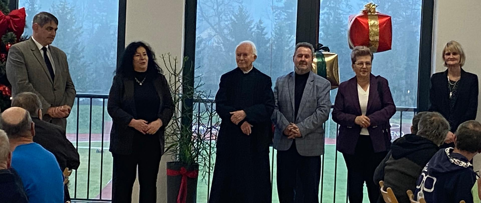 Grupa przedstawicieli władz samorządowych, duchowieństwa oraz organizatorów stoi na sali podczas spotkania wigilijnego. Przed nimi siedzą uczestnicy wydarzenia. W tle widoczne są duże okna, świąteczne dekoracje oraz prezenty, a całość ma uroczysty, świąteczny charakter.