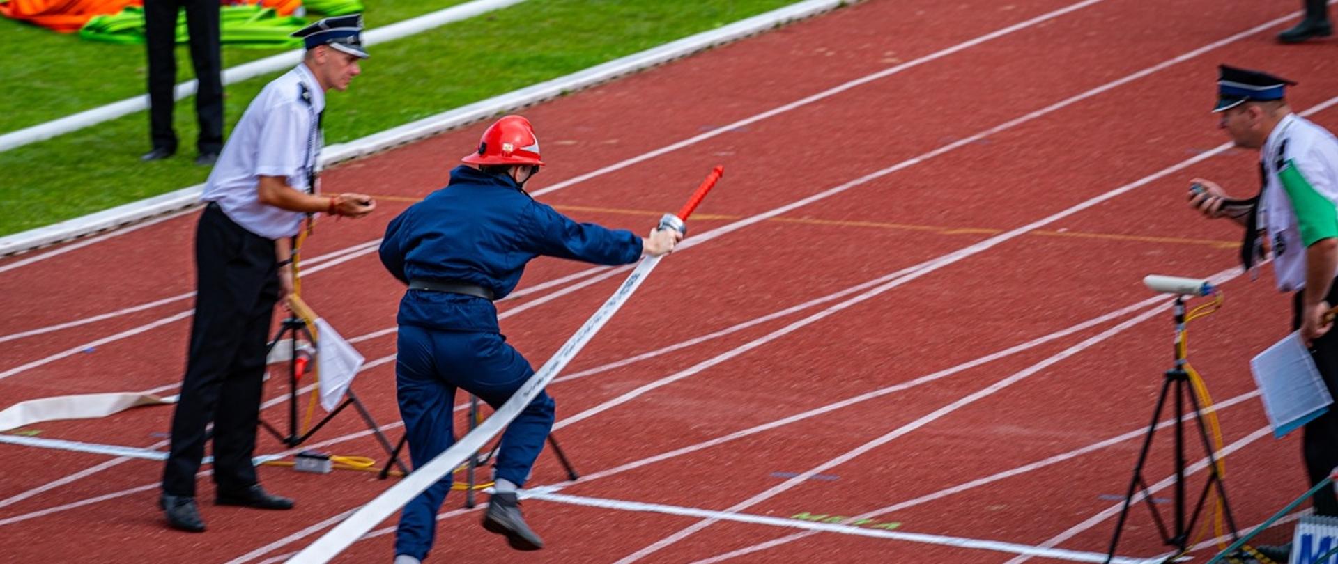 Na bieżni lekkoatletycznej zawodnik ubrany w granatowy kostium i czerwony hełm strażacki dobiega do linii mety z wężem strażackim w prawej ręce. Po obu jego stronach sędziowie odmierzają czas stoperami. W tle murawa ze sprzętem strażackim.