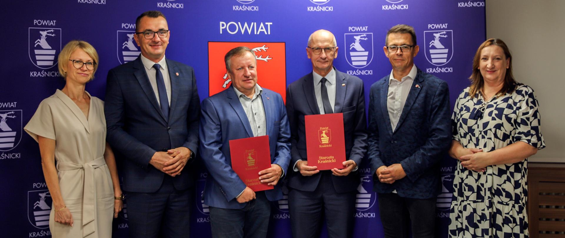 Pamiątkowe zdjęcie z podpisania umowy na realizację inwestycji. Zdjęcie przedstawia sześć osób pozujących do zdjęcia na tle niebieskiej ścianki z wizerunkami herbu powiatu kraśnickiego.