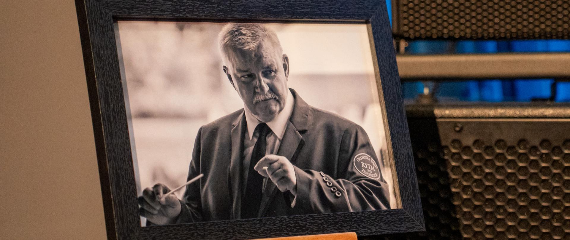 Czarno-białe, oprawione w ramkę zdjęcie mężczyzny w średnim wieku – dyrygenta w garniturze, uchwyconego w trakcie prowadzenia orkiestry, z batutą w dłoni. Fotografia ustawiona jest na drewnianej sztaludze, a w tle widoczny jest fragment sceny z nagłośnieniem.