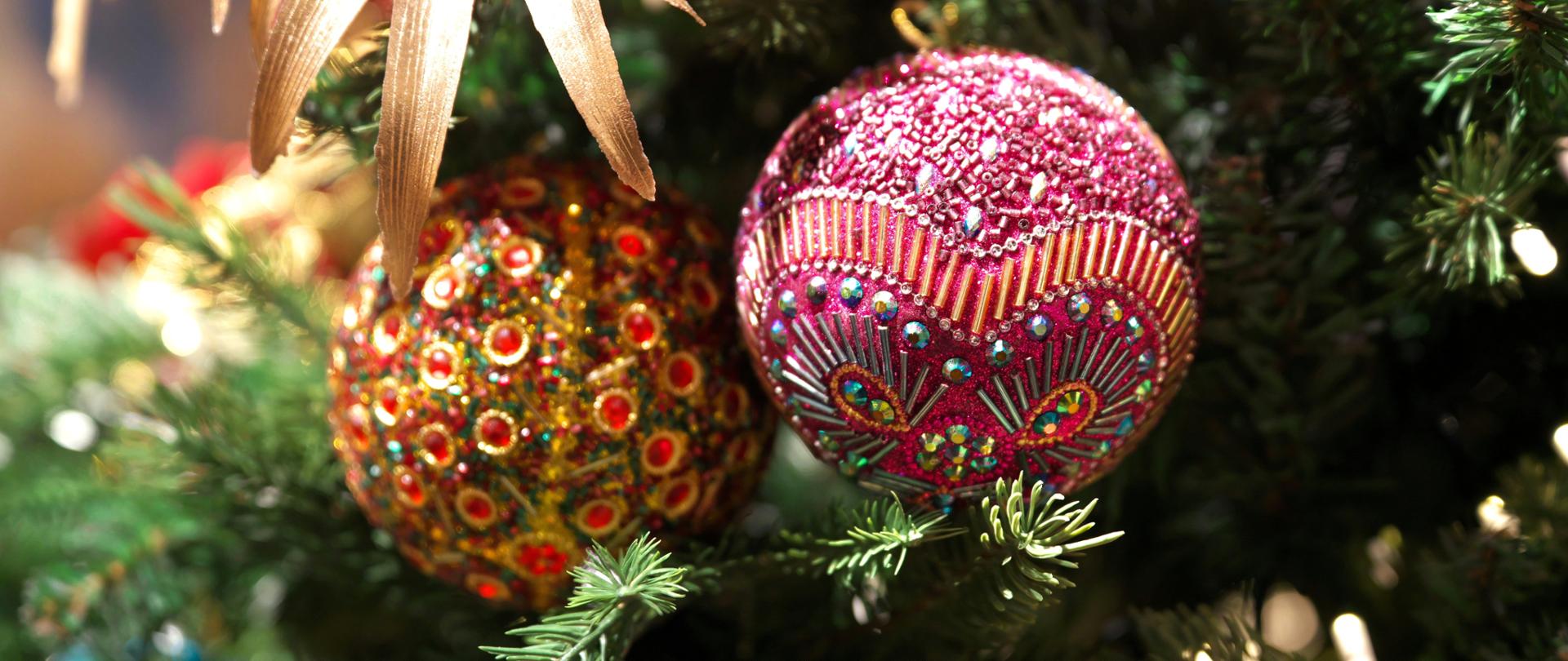 Dwie bogato zdobione bombki choinkowe, zawieszone na zielonych gałązkach choinki. Większa, fioletowo-różowa bombka jest wysadzana błyszczącymi koralikami, ułożonymi w geometryczny wzór. Obok niej znajduje się mniejsza bombka w odcieniach złota i czerwieni. W tle widać złote ozdoby w kształcie liści.