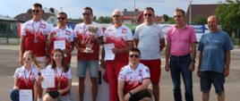 Grupa zawodników modelarskich KM Wierzawice pozuje do zdjęcia z trofeum i dyplomami. Osoby są ubrane w biało-czerwone koszulki z polskim godłem, niektórzy mają medale. Dwie dziewczyny i jeden chłopak klęczą z przodu, pozostali stoją za nimi. W tle widoczne są domy i baner zawodów.