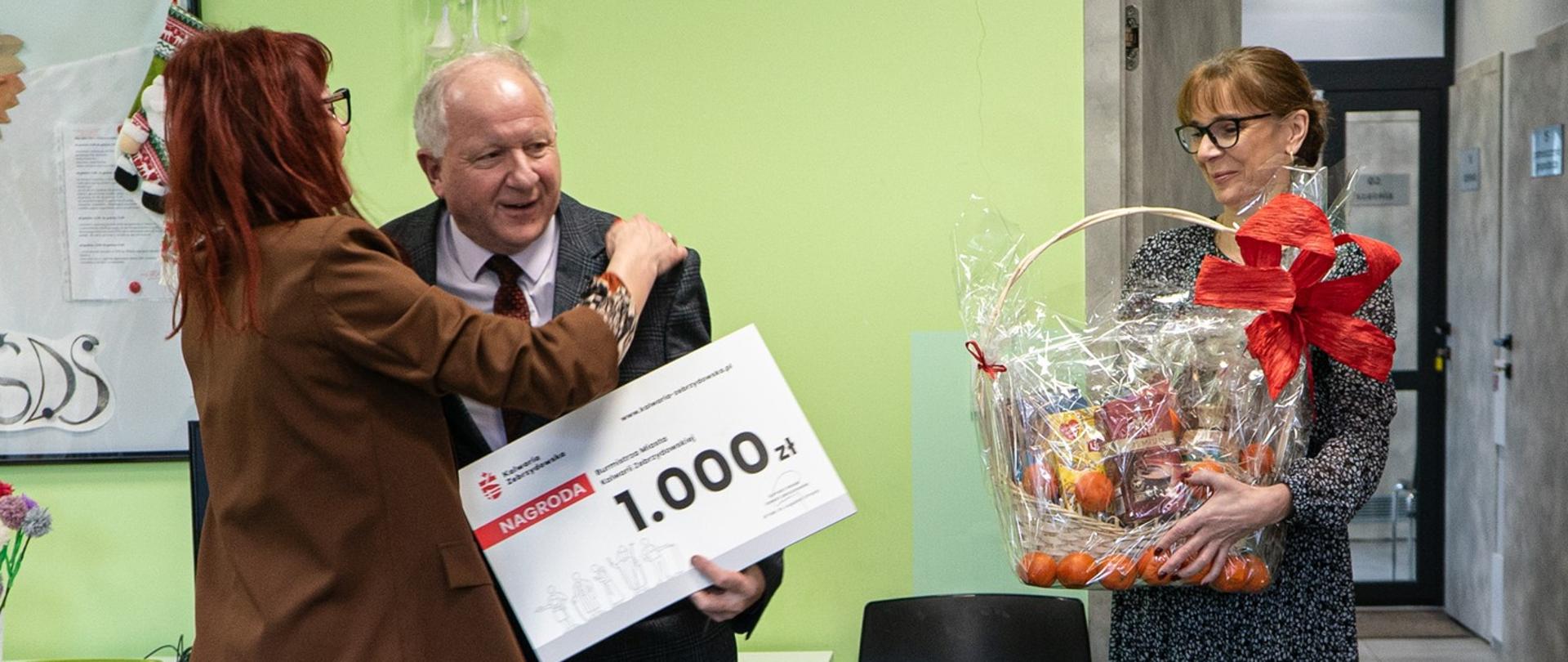 Z lewej strony mężczyzna w garniturze z czekiem o wartości 1000 złotych, po prawej stronie kobieta z koszem słodyczy.